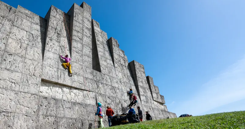 Een aantal mensen klimt tegen een grote, gestructureerde betonnen muur met geometrische vormen en verzamelt zich aan de voet ervan, onder een helderblauwe hemel met felgroen gras op de voorgrond.